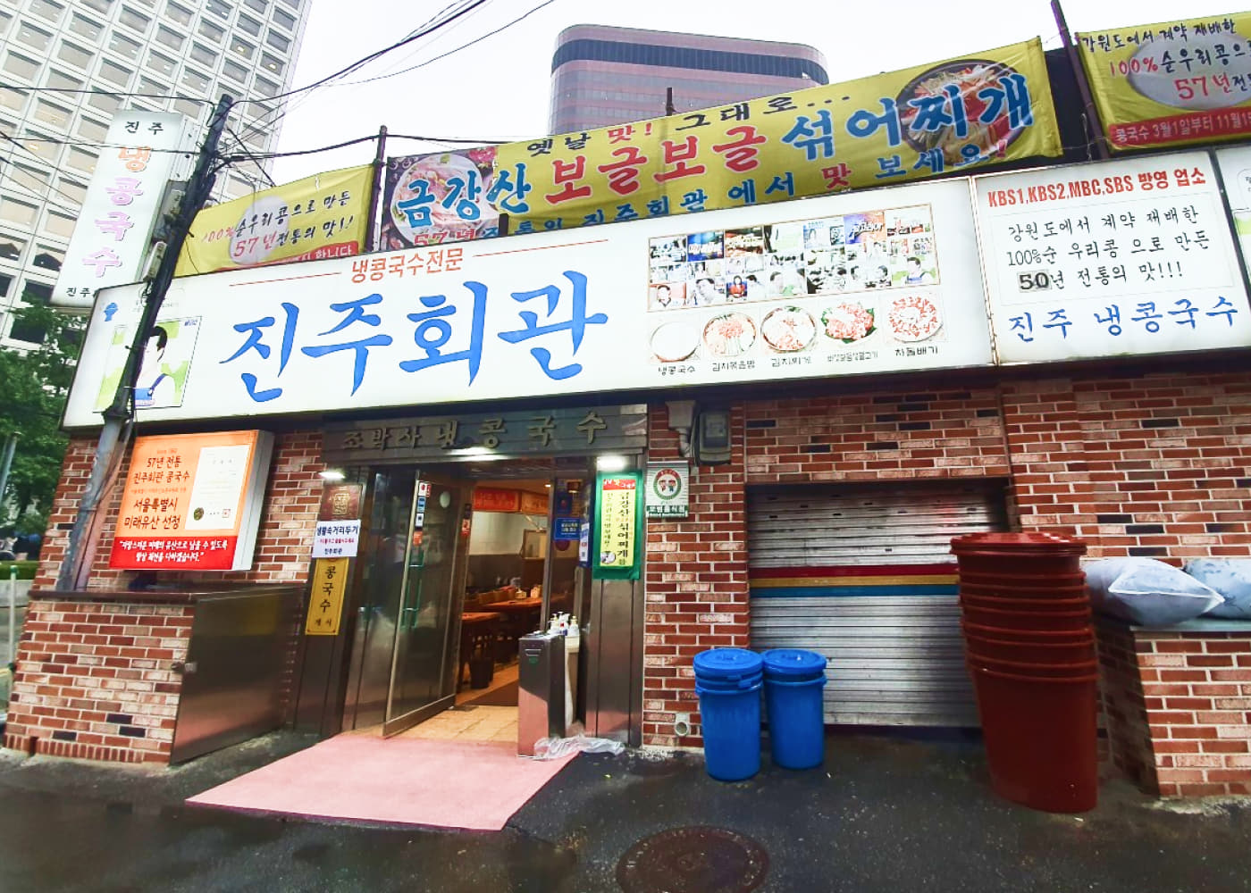 붉은 벽돌 외관과 큰 진주회관 간판, 비에 젖은 길바닥이 함께 보이는 시청 진주회관 정면