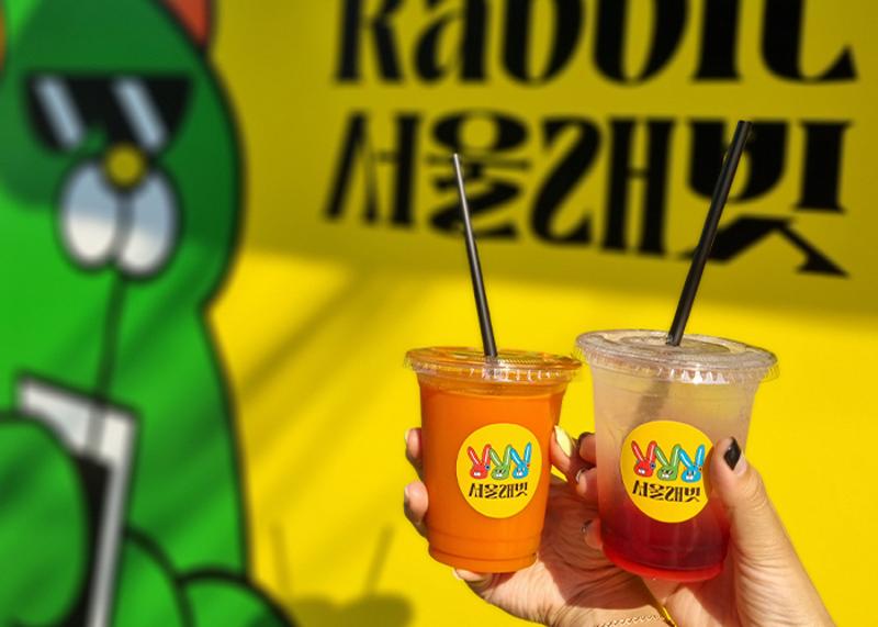 서울 래빗의 당근 음료 두 잔을 노란 벽과 토끼 캐릭터 앞에서 들고 있는 장면